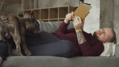 hombre acostado en el sofá con su perro bulldog mientras lee un libro