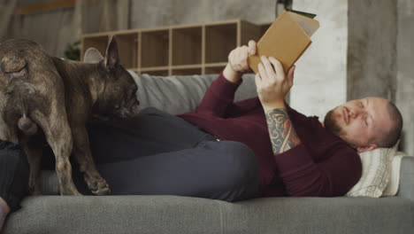 man lying on the sofa with his bulldog dog while reading a book