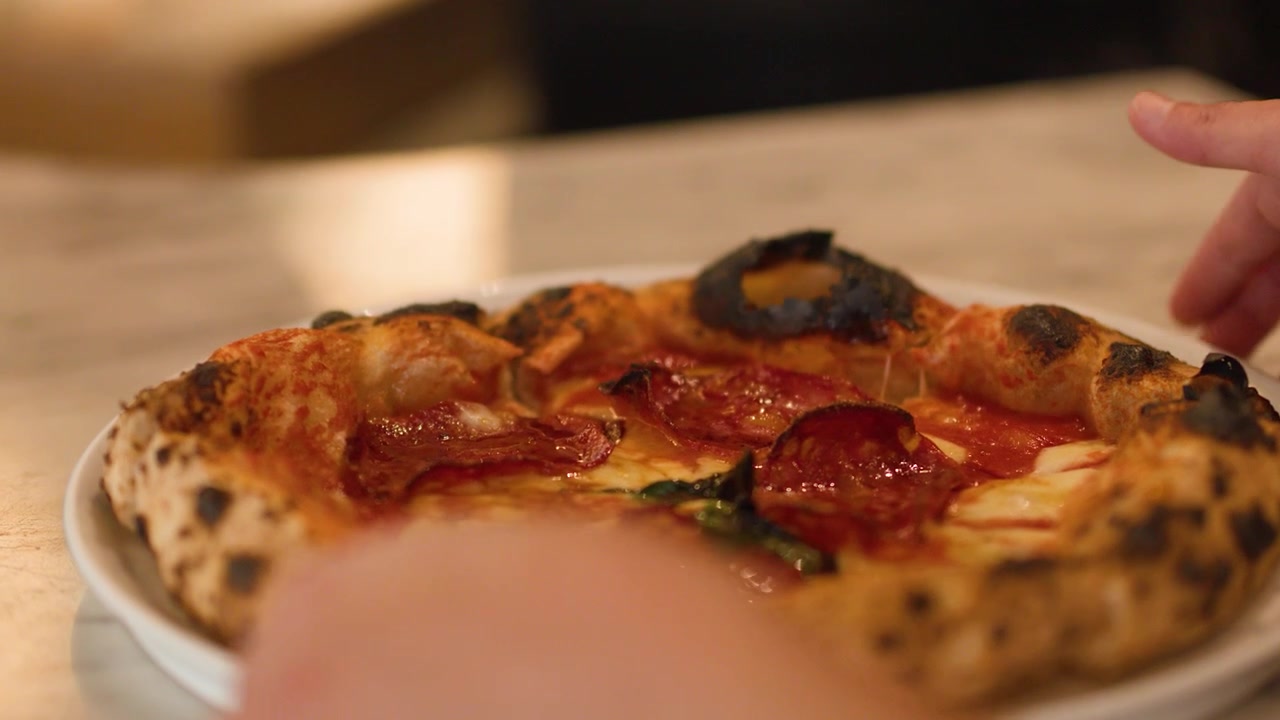 Primo piano di mani che sollevano una pizza al salame piccante con crosta di pasta madre da un tavolo di marmo di un ristorante.