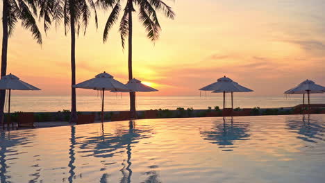 Perfect-red-sun-sunset-view-from-tropical-hotel-resort-lounge-near-infinity-pool,-beach-umbrellas,-and-coconut-palms-in-the-foreground