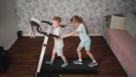 boy in white shirt grips treadmill handle for support while girl behind him stretches arms toward same handle during shared fitness activity in cozy room with wooden floor