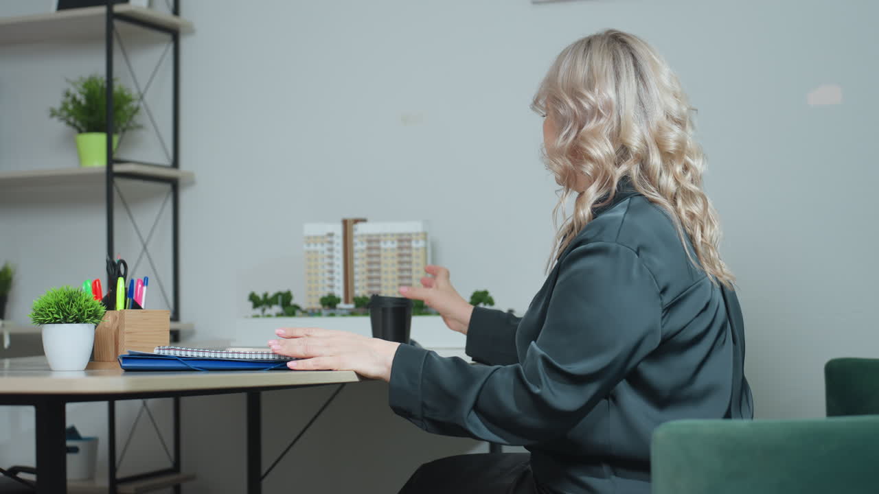 Rear view of woman seated in modern office opening laptop and adjusting coffee cup on clean work desk, preparing for productive day with notebooks, pens, and real estate model