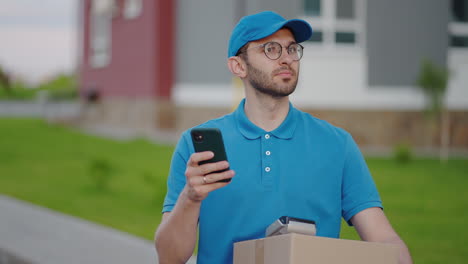 the postman with glasses carries the parcel and looks at the delivery address via mobile phone. search for the address of the delivery customer. delivery guy with a cap and a box