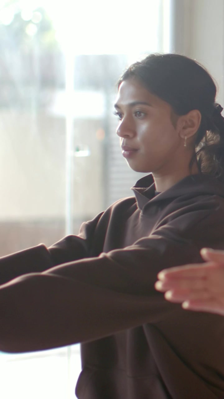 Vertical video: Mirroring other hand, woman in dark brown hoodie extending arms by glass doors