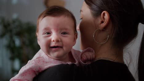 retrato en primer plano de una pequeña niña en un traje rosa sentada en los brazos de su madre y mirando por encima de su hombro posando en un apartamento moderno