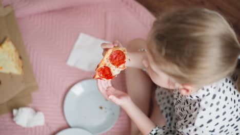 overhead view of young girl in spotted dress holding pepperoni pizza slice above empty plates while sitting on pink blanket beside open pizza box