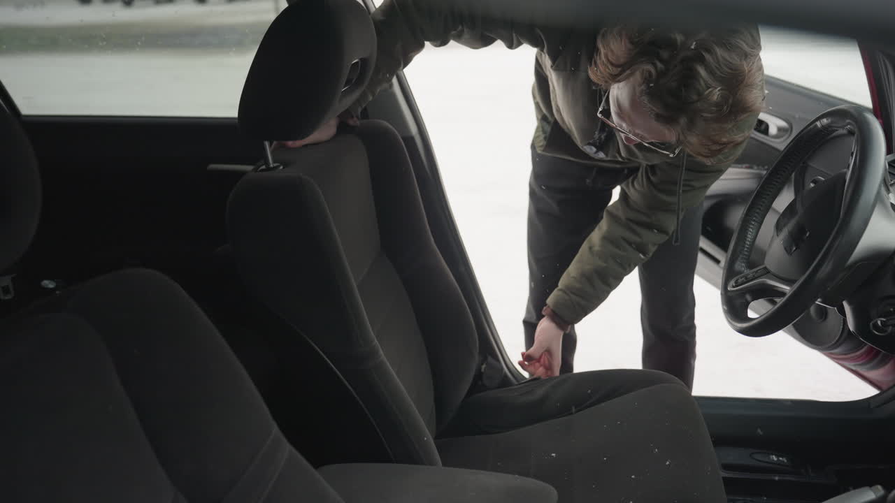 man adjusts front seat of parked car interior during snowy day with another vehicle seen through open door window in blurred background, emphasizing winter maintenance activity in cold weather