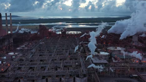 Drone-shot-orbiting-coal-and-metal-factory-at-dawn,-large-steam-smoke-stacks-operating