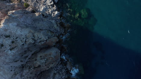 Slow-Overhead-Top-Down-Aerial-along-Generic-Rocky-Coast-with-Turquoise-Water-and-Waves-Crashing-on-Rocks-on-Greek-Island-at-Sunset