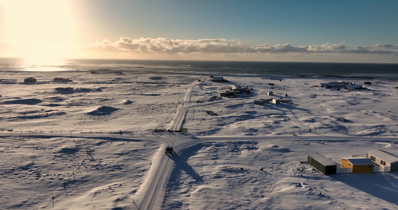 Aerial view of a lone 4x4 turning through a snow-covered coastal crossroads near Strandarkirkja, with low Arctic sun, scattered buildings, and open Atlantic horizon.