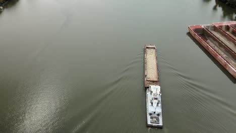 aerial drone footage of towboat guiding cargo barge along calm kanawha river surrounded by forested hills, industrial docks, and quiet roads in scenic west virginia, united states
