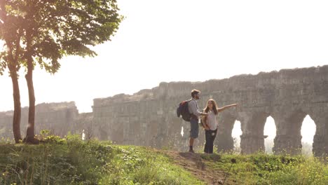 joven pareja encantadora mochileros turistas leyendo mapa señalando direcciones acueducto romano arcos en parco degli aquedotti parque ruinas en roma en romántico amanecer brumoso con guitarra y saco de dormir cámara lenta