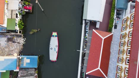 Aerial-Birds-Eye-View-Flying-Along-Khlong-Bangkok-Yai-Canel-Waterways-With-Boat-Sailing-Below