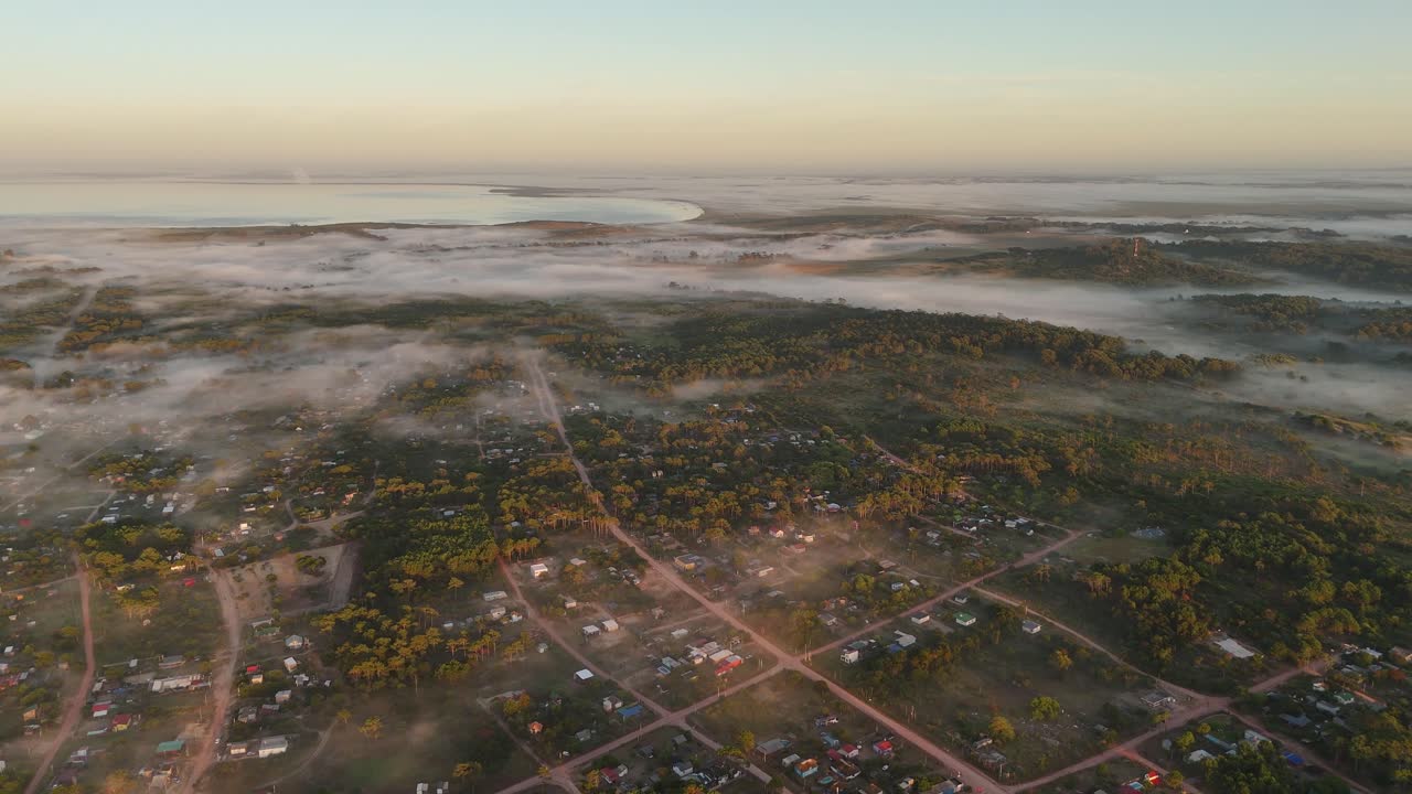 A breathtaking aerial view of coastal town Punta del Diablo, gently cloaked in a thick layer of fog at dawn, with Black Lagoon visible in background.
