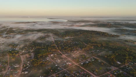 a breathtaking aerial view of coastal town punta del diablo, gently cloaked in a thick layer of fog at dawn, with black lagoon visible in background.
