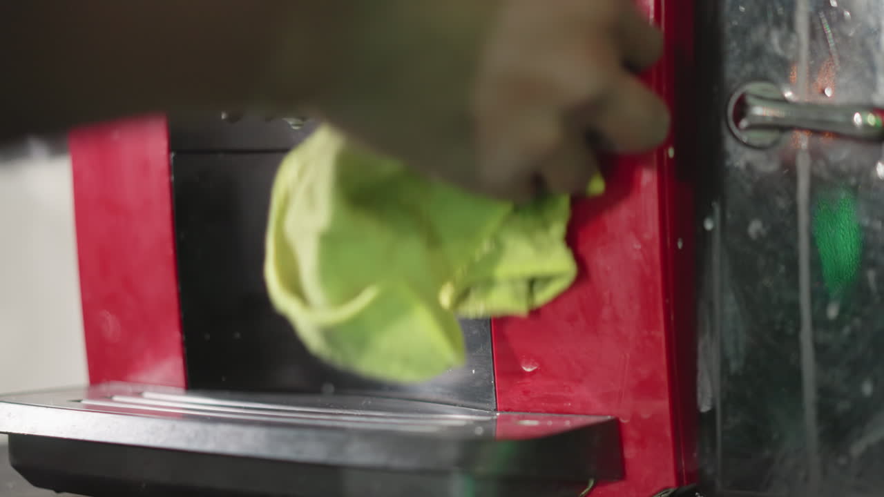 Close-up of person cleaning coffee machine with green cloth, hand wiping black surface of appliance, focusing on detail and maintenance, showing effort to keep kitchen equipment clean