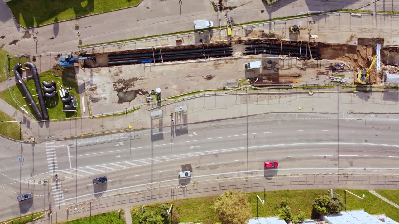 Aerial bird's eye view above sewer pipe installation at construction site as pedestrians and cars pass by on roadway, Purvciems construction site in Latvia