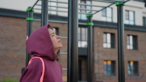 self driven woman in hoodie carrying bag pauses before urban exercise bars against high rise backdrop on crisp morning stroll embodying determination and active lifestyle in outdoor fitness setting