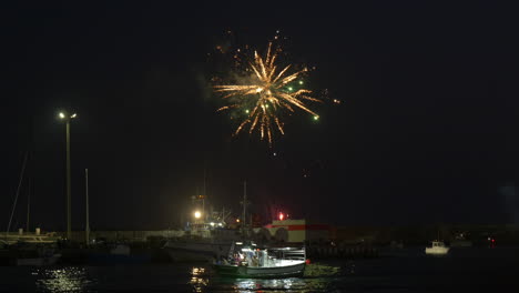 fireworks light up the night sky over the port of barbate, celebrating the day of the procession of the virgin of carmen. the lights are reflected on the water, with a boat passing by