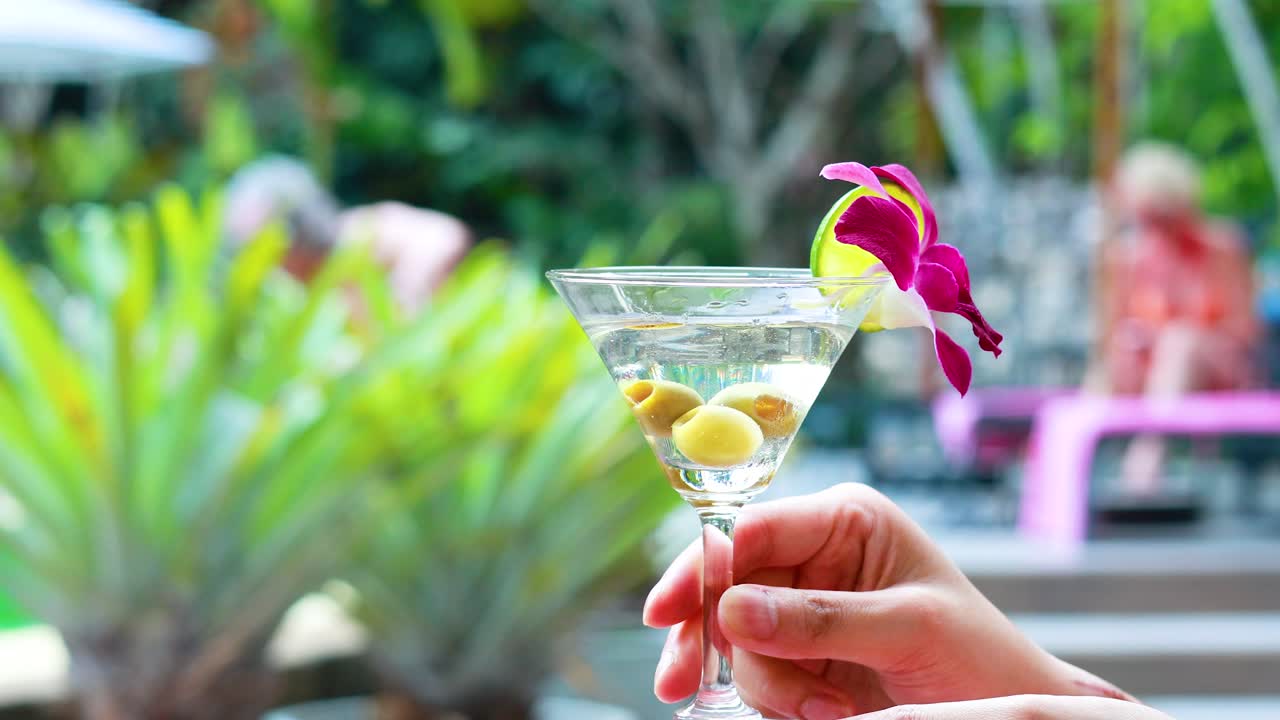 손은 오키디 장식으로 마르티니를 들고, 활기찬 수영장 배경을 배경으로 편안한 휴가 분위기를 포착합니다.