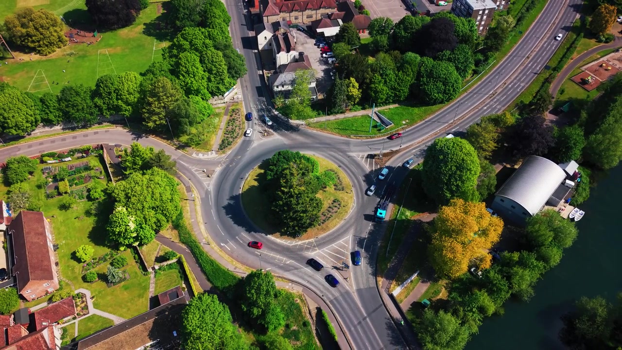 Top down aerial drone shot of a road roundabout in Salisbury, Wiltshire, showing cars moving around the junction, surrounding trees, green space, footpaths and urban roads on a sunny day.