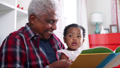 grandfather sitting and reading book with baby granddaughter at home