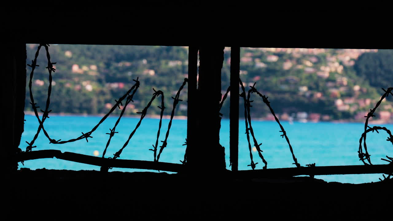 Close up of barbed wire loops along a small window with a blurred view of the sea and a town