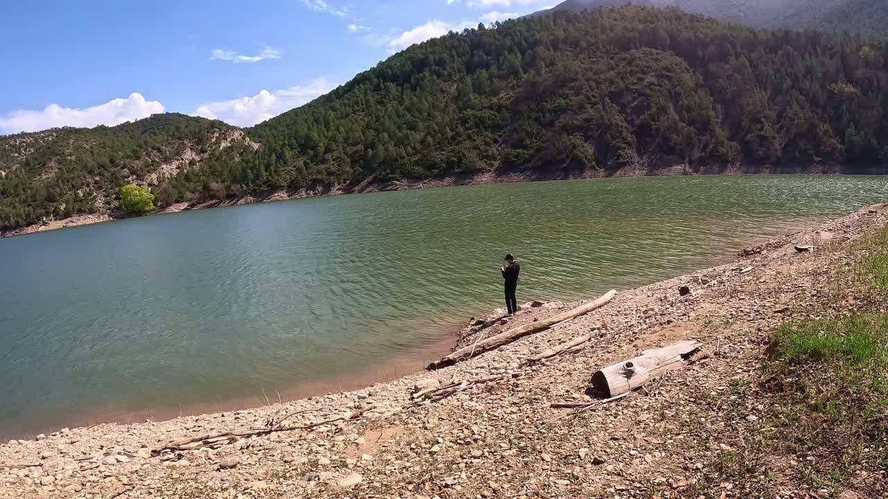 A lone fisherman standing on a rocky shoreline, casting his line into a serene lake surrounded by lush forested hills under a clear blue sky