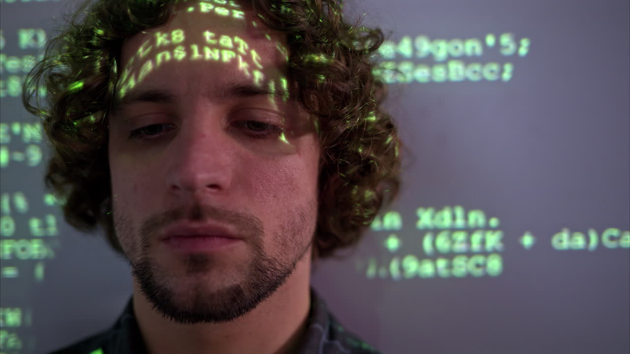 Man with curly hair in front of a projection of green computer code, illuminating on his face