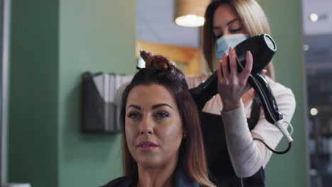 Female-hairdresser-wearing-face-mask-blow-drying-hair-of-female-customer-at-hair-salon