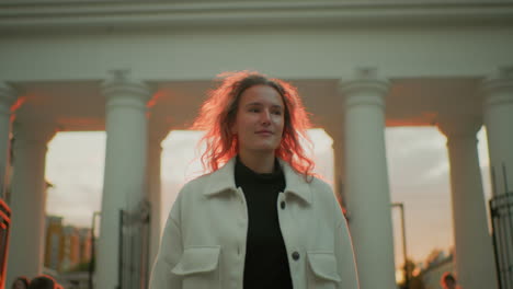young lady wearing long white coat confidently strolling outdoors with soft sunlight illuminating her hair, standing against background of white pillars and open gates during warm golden hour evening