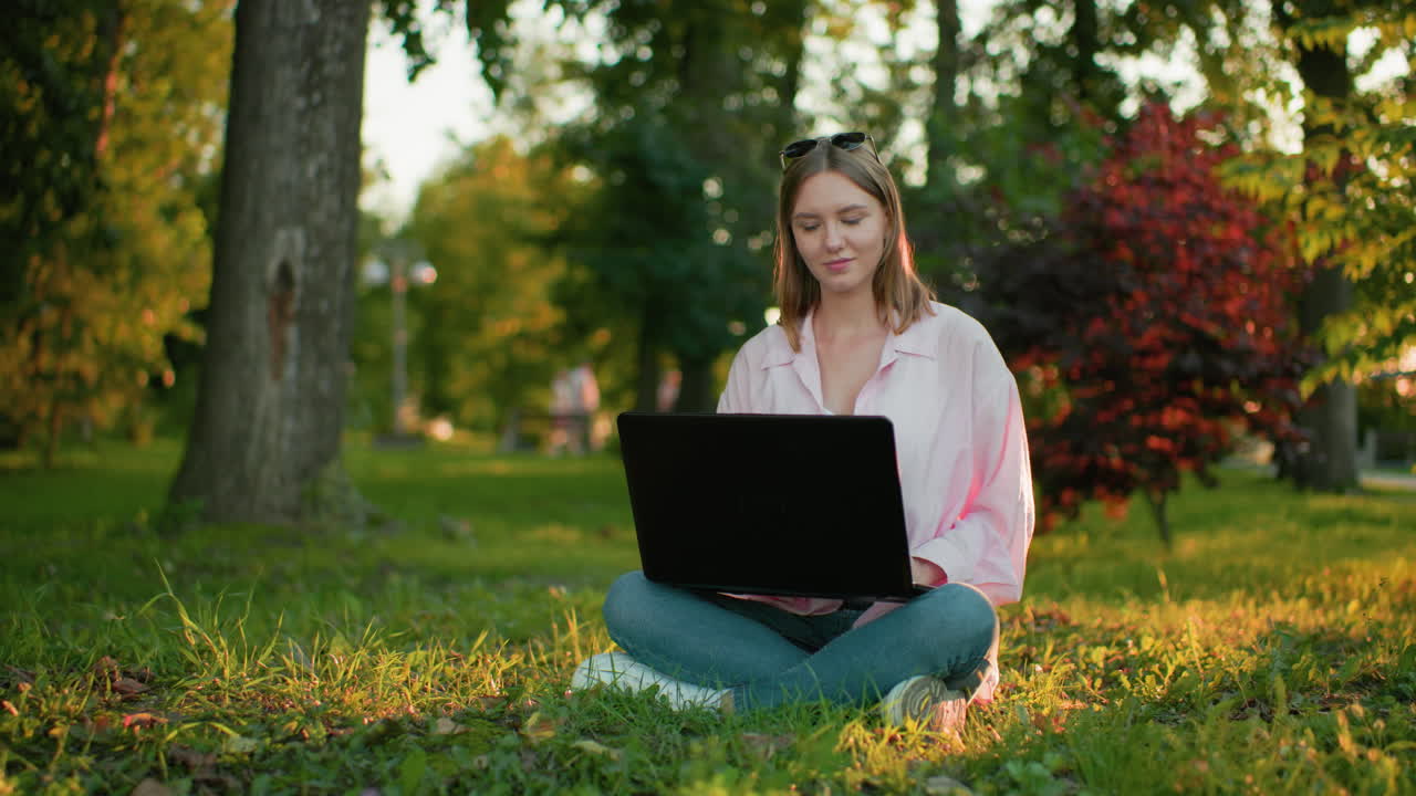 Freelancer in pink top with glasses resting on her head smiles warmly and continues typing on her laptop while sitting outdoors, trees and people walking appear in background with natural lighting