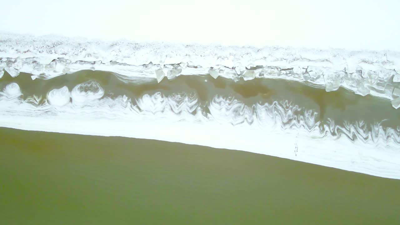 Drone flies along frozen winter coastline showing snow covered beach, floating ice chunks, and cold green sea water