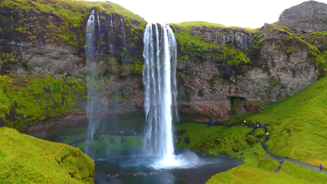 fotografía de un avión no tripulado de una gran cascada en el sur de islandia con vegetación alrededor, un rayo de sol y turistas
