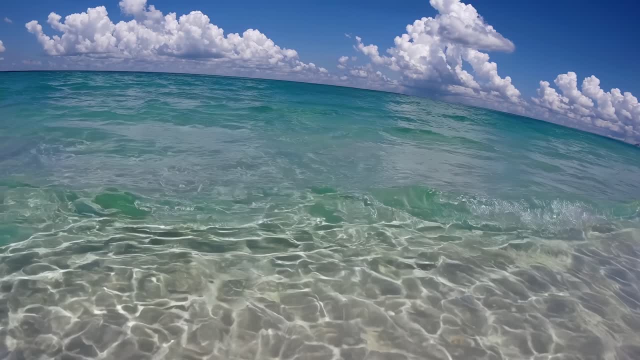 Videobild i låg vinkel av havsvågor som försiktigt sköljer över stranden under en klarblå himmel med fluffiga moln.