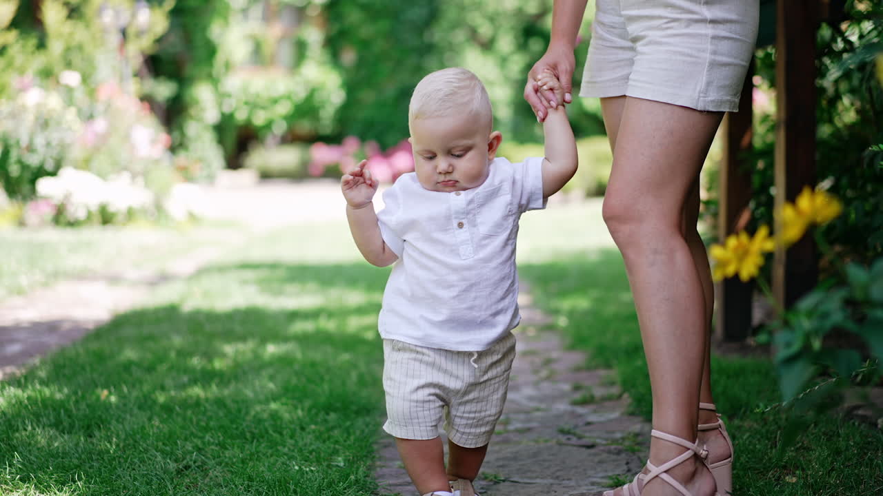 Unrecognized mother stands outdoors holding her lovely baby by the hand. Cute blond child learns to walk.