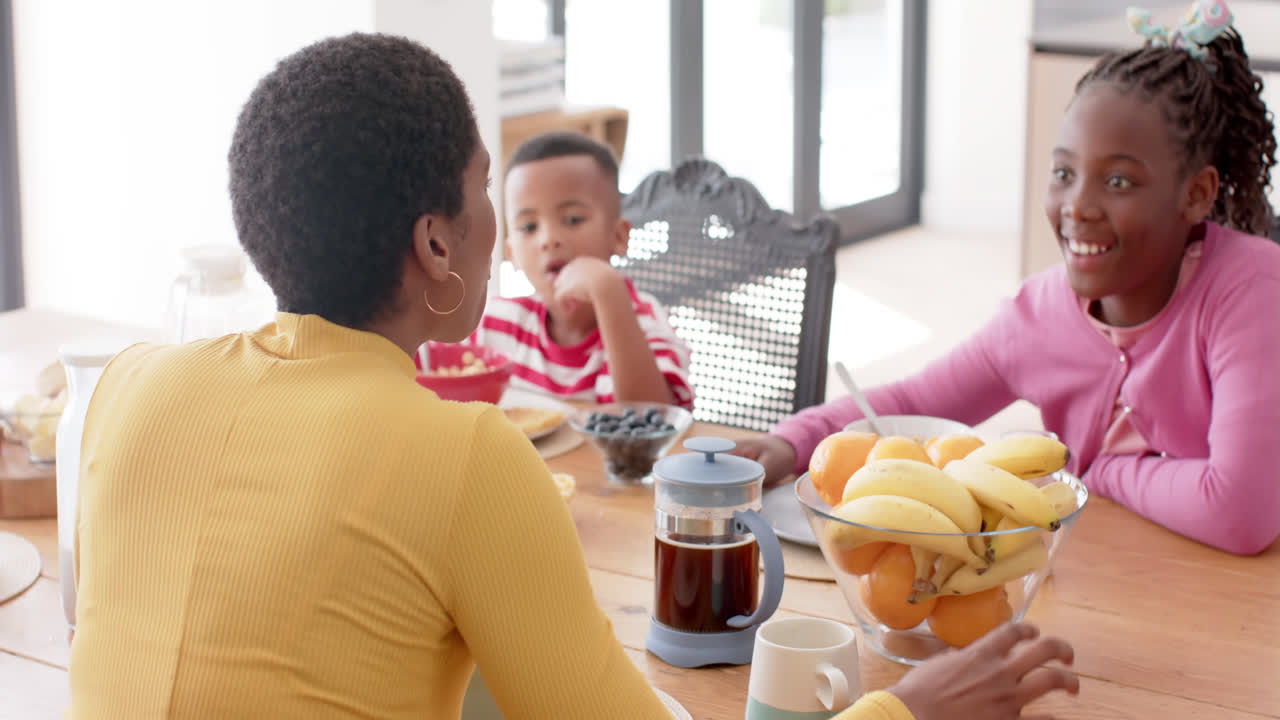 madre afroamericana con hijo e hija comiendo el desayuno en la mesa en la cocina, cámara lenta