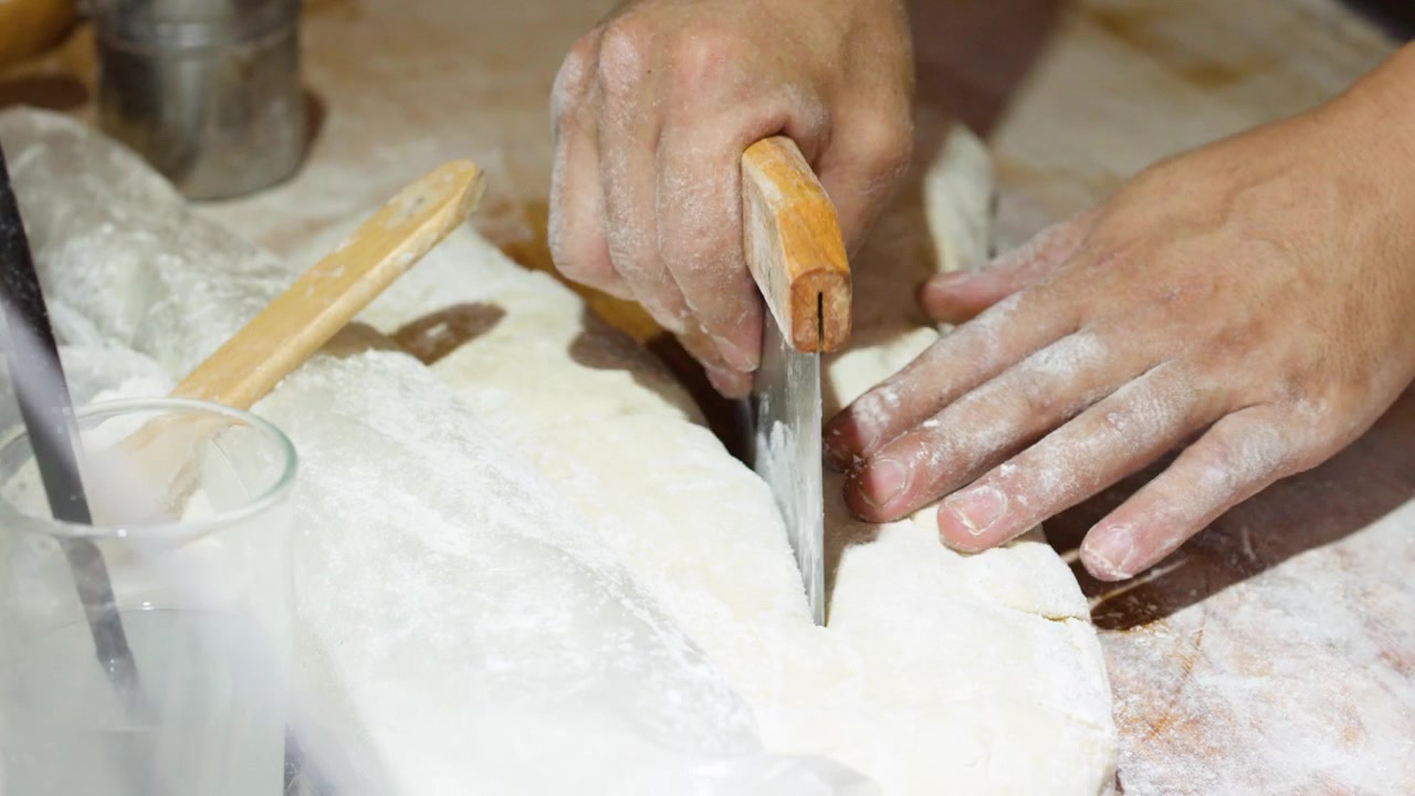 Primo piano delle mani di un fornaio che tagliano e modellano l'impasto di pane fresco con un raschietto da banco in metallo