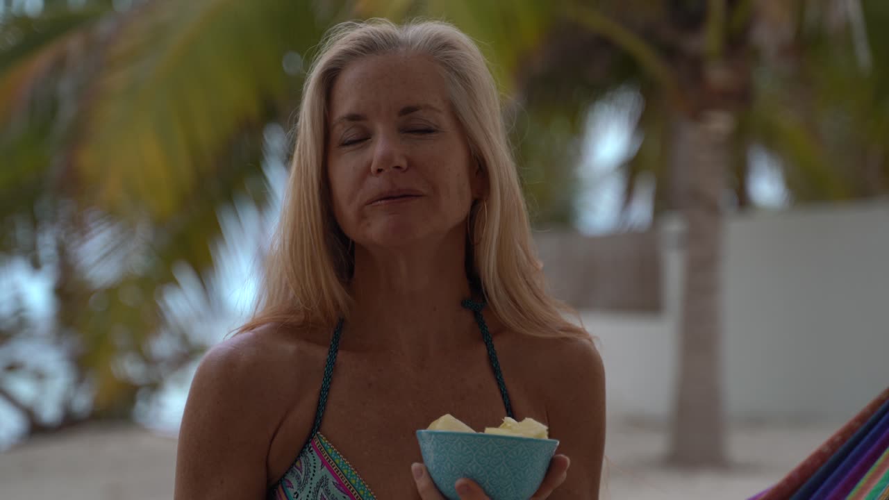 리조트에서 해먹에 앉아 비키니 입은 꽤 성숙한 여성의 근접 촬영, 파인애플 한 조각을 맛보고 카메라에서 미소