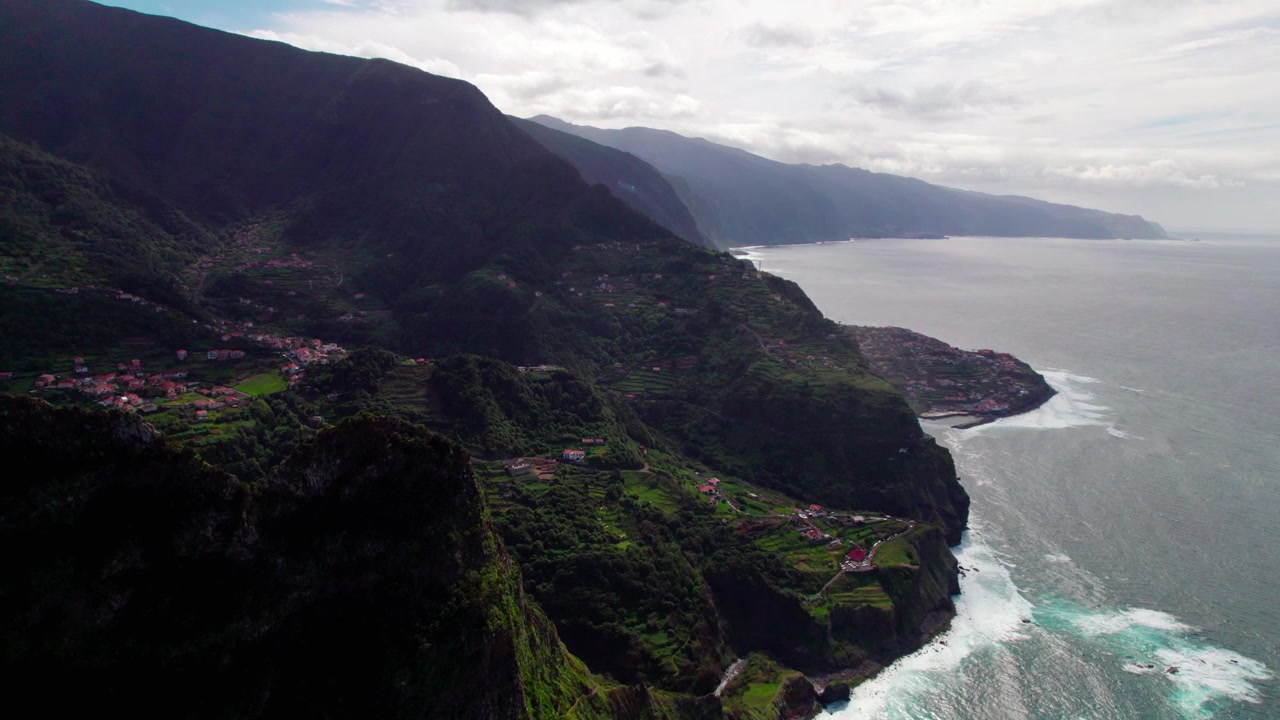 Una spettacolare vista aerea mostra ripide scogliere costiere, paesaggi terrazzati verdi, case sparse sul fianco della collina e onde che si infrangono sulla costa frastagliata sotto un cielo nuvoloso - Ripresa aerea con drone