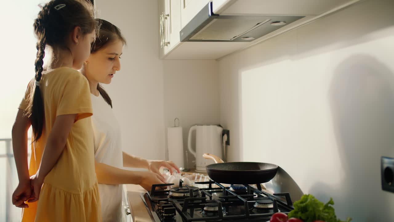 una mujer morena en una camiseta blanca junto con su hija pequeña en un vestido amarillo le dice cómo usar correctamente la estufa y cocinar comida en la cocina en un apartamento moderno