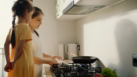 A-brunette-woman-in-a-white-T-shirt-together-with-her-little-daughter-in-a-yellow-dress-tells-her-how-to-properly-use-the-stove-and-cook-food-in-the-kitchen-in-a-modern-apartment