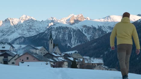 Young-caucasian-man-in-yellow-jacket-and-beanie-walking-and-stopping-to-see-view-of-small-mountain-village-and-church-in-Guarda,-Switzerland