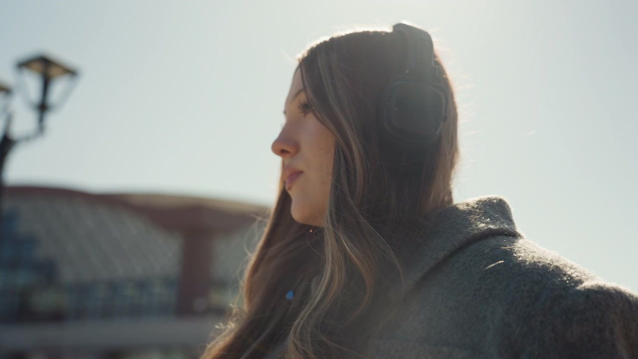 Piazza luminosa con donna pensierosa. Serena signora asiatica alla luce del sole tra gli archi del mercato con le cuffie. Momento tranquillo catturato di una donna asiatica in una piazza illuminata dal sole che indossa un cappotto grande e le cuffie.