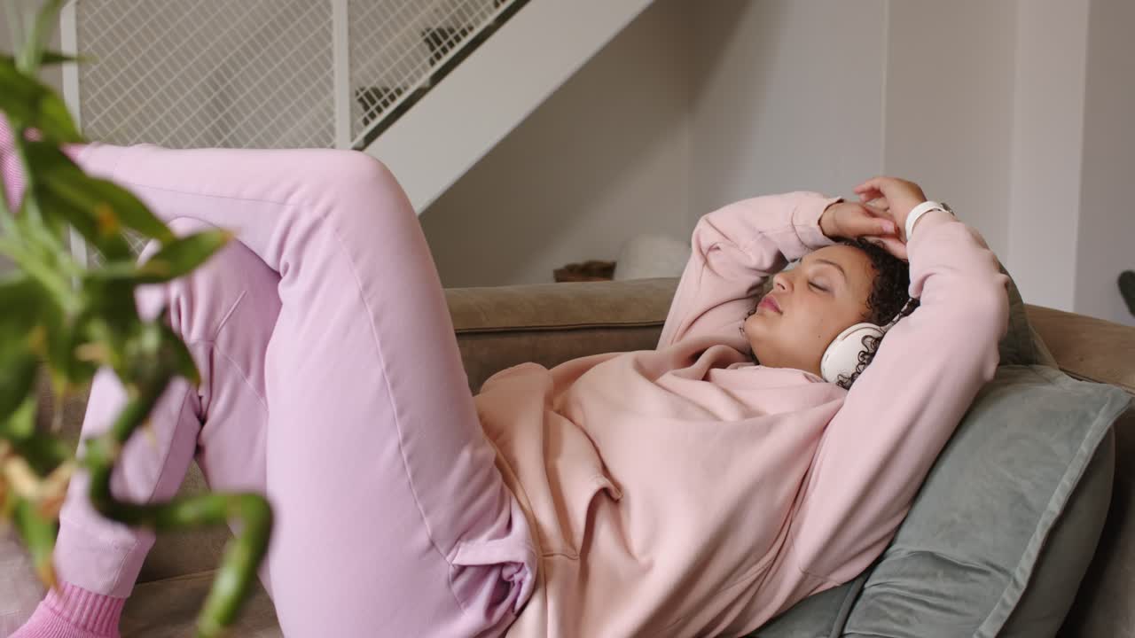 African American woman reclining on couch in living room adjusting white headphones while listening