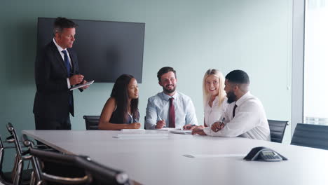 Group-Meeting-Around-Table-At-Graduate-Recruitment-Assessment-Day-And-Collaborating-On-Task-Whilst-Being-Observed-By-Mature-Businessman