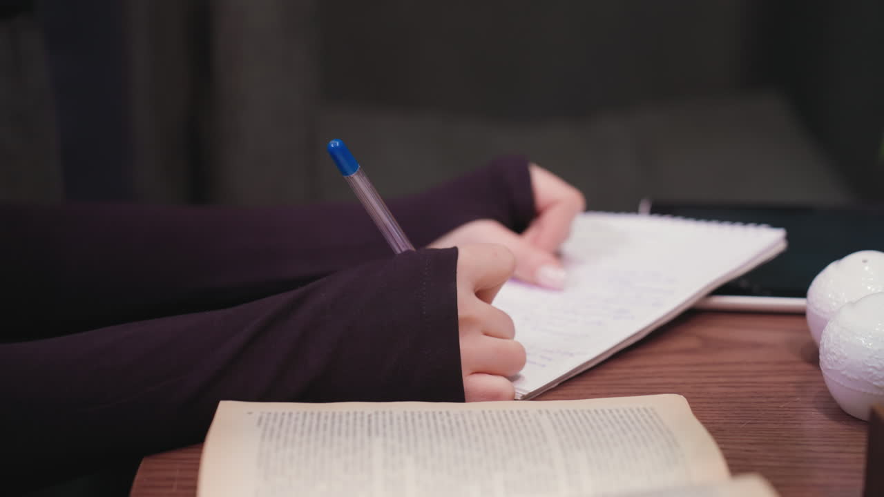 Close up of woman hand holding blue pen while writing in notebook on brown wooden table with blurred open book and white decoration in foreground, creating focused and calm study atmosphere