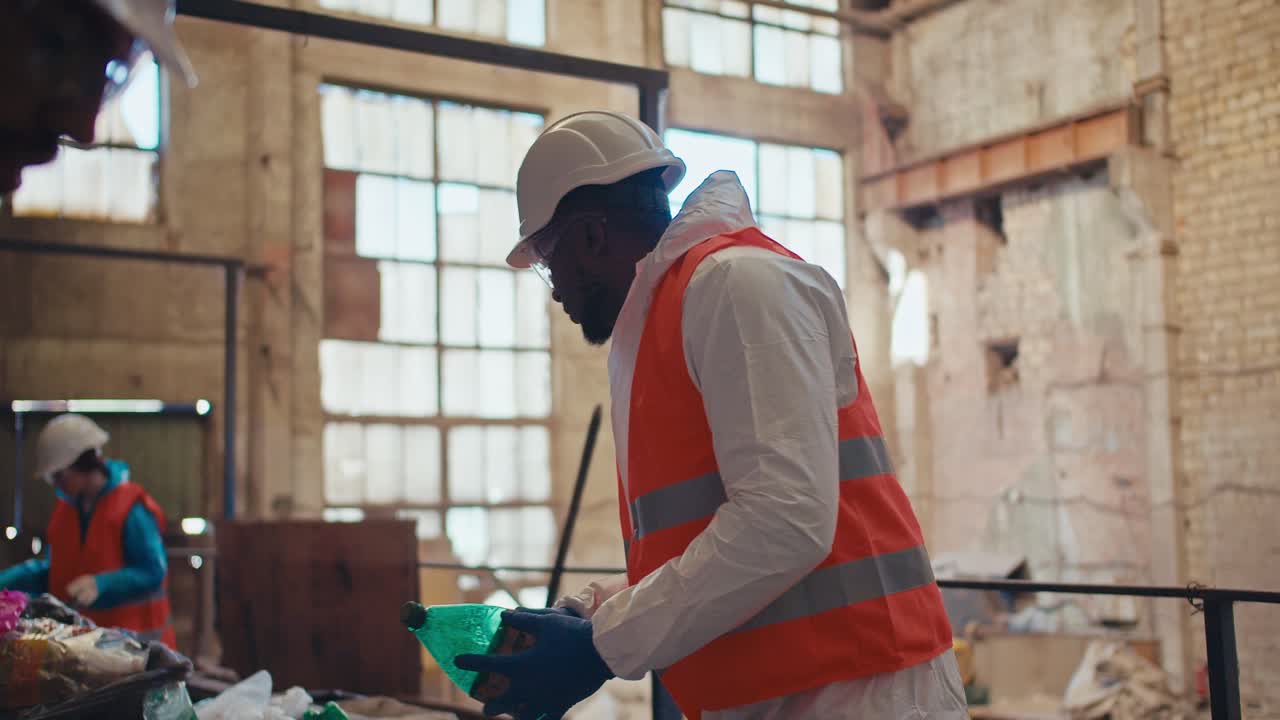 A man with Black skin in a special white protective uniform and an orange vest sorts plastic at an old waste processing plant. A man with Black skin is an employee of a huge waste recycling plant