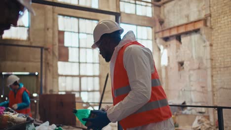 Ein-Mann-Mit-Schwarzer-Hautfarbe-In-Einer-Speziellen-Weißen-Schutzuniform-Und-Einer-Orangefarbenen-Weste-Sortiert-Plastik-In-Einer-Alten-Müllverarbeitungsanlage.-Ein-Mann-Mit-Schwarzer-Hautfarbe-Ist-Angestellter-Einer-Riesigen-Müllrecyclinganlage