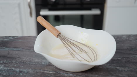 whisk resting in baking mixture inside white bowl on countertop, kitchen background features modern cabinets, oven, and clean workspace, creating a homey baking atmosphere